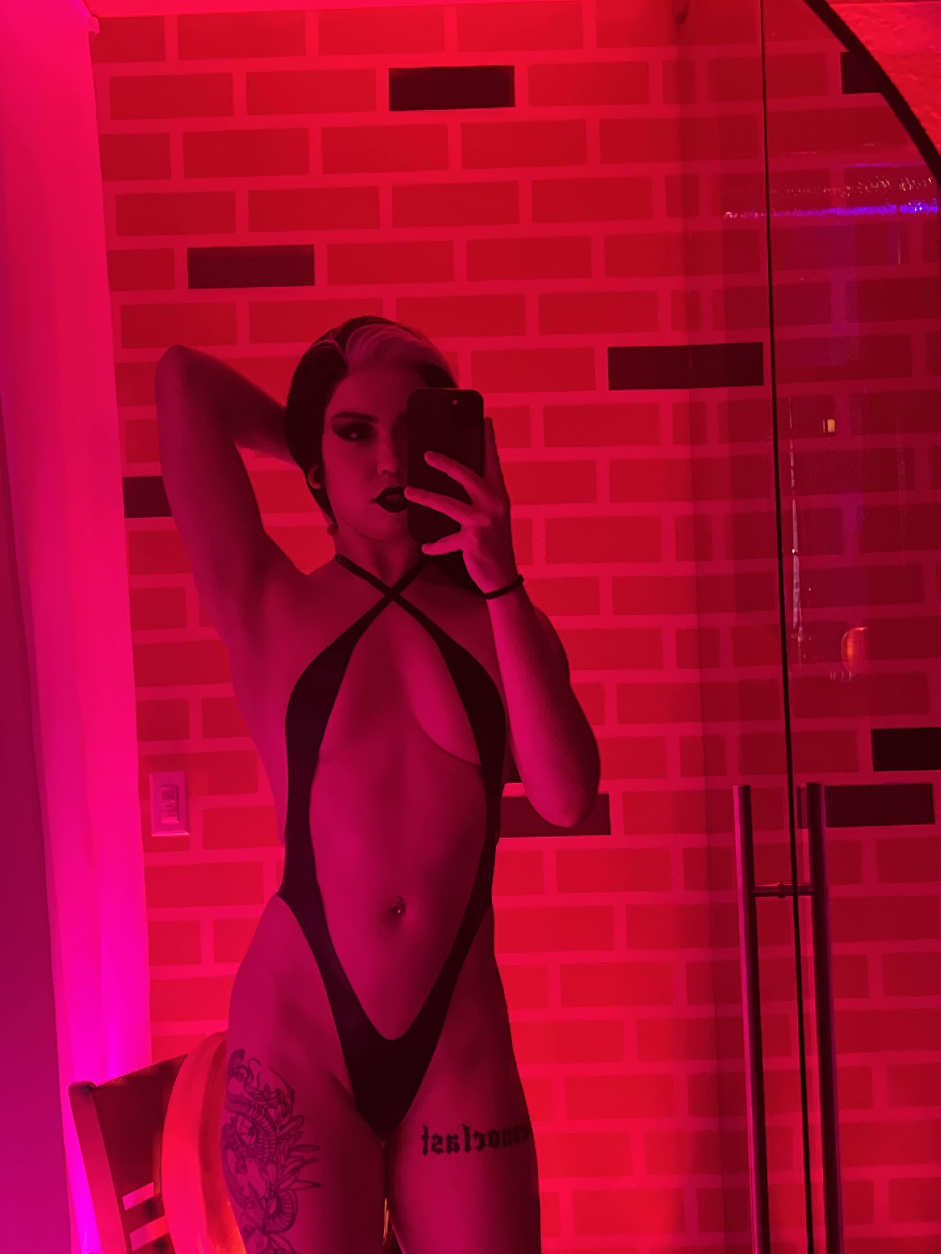 a selfie of bex in a red room wearing a black latex one piece